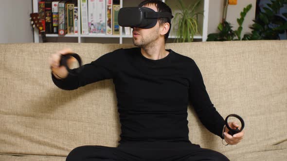 A bearded man wearing a VR helmet and black clothes sits on a couch. In his hands are controllers