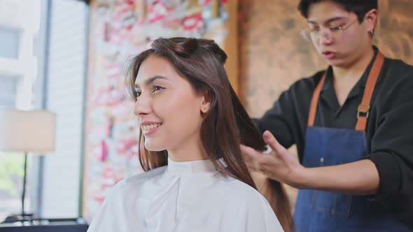 Asian professional male hairstylist combing and using scissors cutting woman's hair in beauty salon.