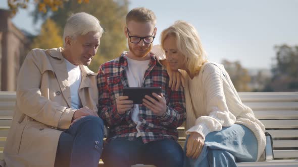 Senior Parents and Adult Son Sitting on Bench Using Digital Tablet Outdoors