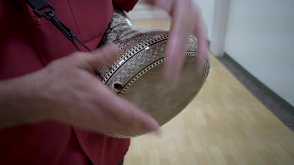 Tight shot of man playing pearl encrusted dumbeck with a shoulder strap in a wood floored room with