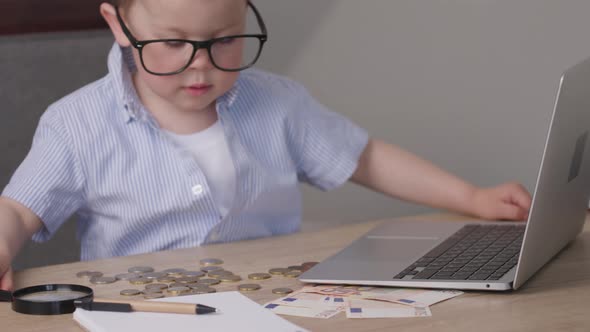 Kid Boy Child in Blue Shirt Typing Laptop Money Euro Banknotes Coins on Table