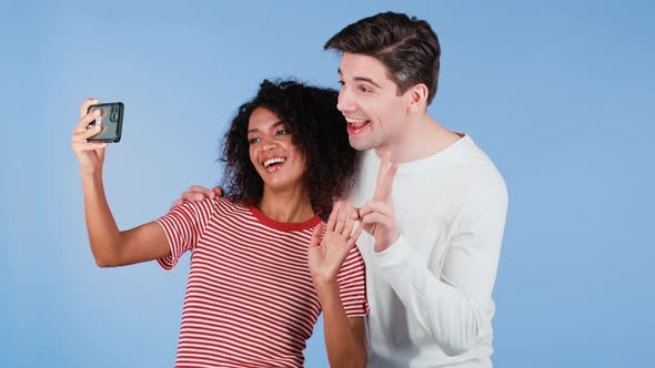 Young Interracial Couple Making Selfie with Smartphone on Blue Studio Background