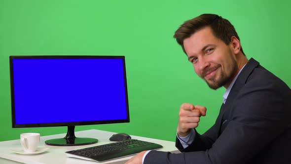 A Young Businessman Sits in Front of a Computer and Points at the Camera - Green Screen Studio