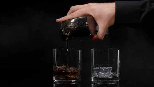 Barman Pouring Whiskey Cognac Brandy From Bottle Into Glasses with Ice Cubes on Black Background