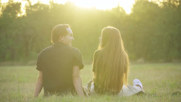 Relaxed Caucasian Adolescent Couple Sitting in Sunbeam on Summer Meadow Talking and Smiling