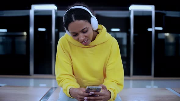 Young Girl Looks at Her Phone Listens Music in Headphones Sitting at Station