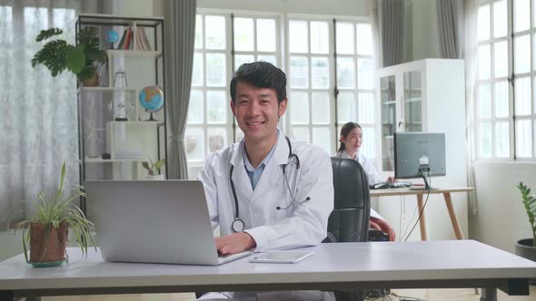 Young Asian Man Doctor Is Using Laptop Computer, He Turns And Smiles To Camera In Workplace