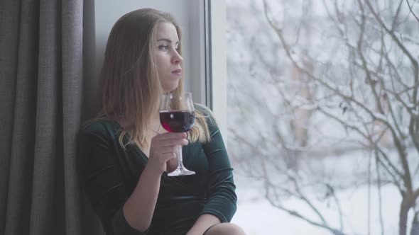 Thoughtful Young Caucasian Woman Looking Out the Window and Drinking Red Wine From Glass. Lonely