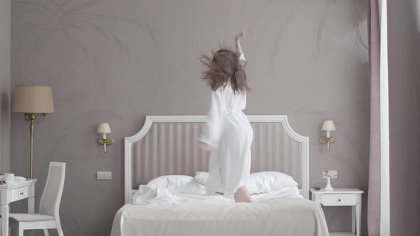 Funny Young Caucasian Woman Jumping on Bed in Hotel Room. Wide Shot Portrait of Cheerful Brunette