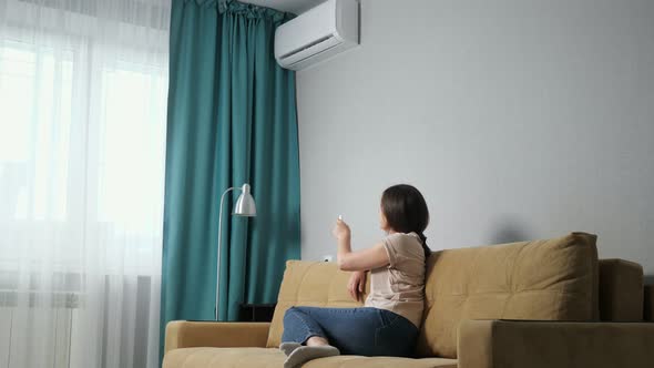 Side View of a Brunette Woman Adjusts the Air Conditioner While Sitting on the Sofa