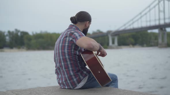Back View of Absorbed Caucasian Guitarist Playing on River Bank with Blurred Bridge at the