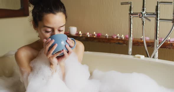 Relaxed biracial woman lying in bath with foam and drinking coffee