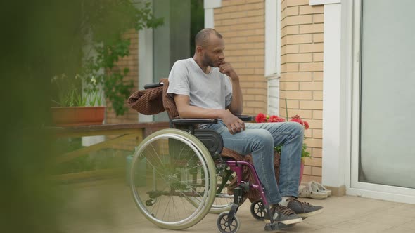 Side View of Thoughtful Sad Disabled African American Man in Wheelchair on Porch in Backyard Garden