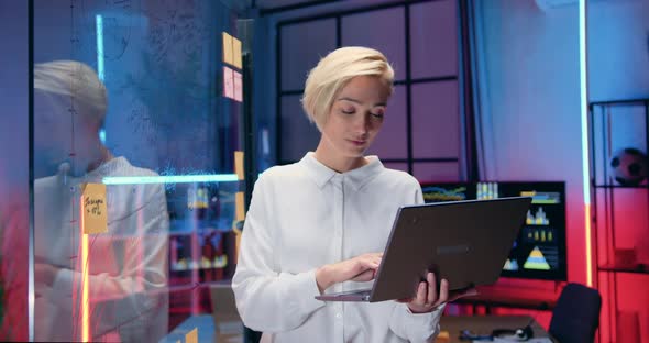 Businesswoman Typing on Laptop, Standing Near Glass Wall in Evening Office