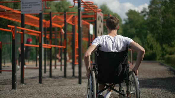 Back View Young Man Rolling Wheelchair on Sports Ground in Spring Summer Park Looking Around