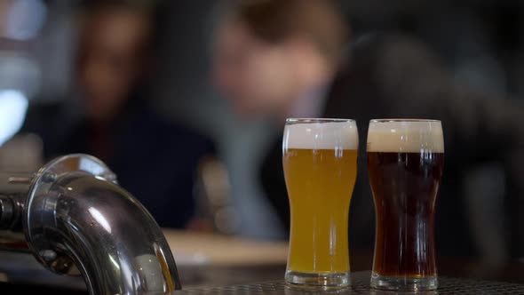 Closeup Pale and Dark Lager in Glasses on Bar Counter with Blurred Caucasian and African American