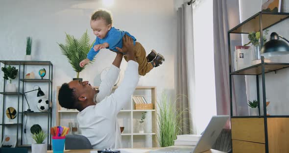 African American Father Sitting on the Chair in Cozy Room and Holding His Cute Cheerful Small Son