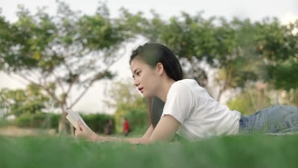 A beautiful Asian woman relaxed reading a book while lying on the floor grass outdoors.