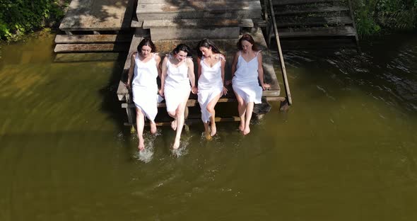 Girls In White Dresses Sit On The Pier And Splash In The Water