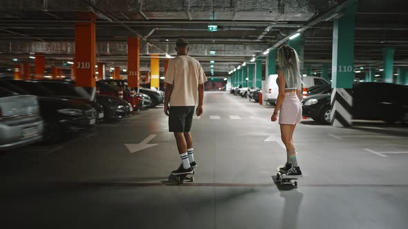 Back View Follow Shot of Young Trendy Diverse Couple Riding Skateboards at Urban Underground Parking