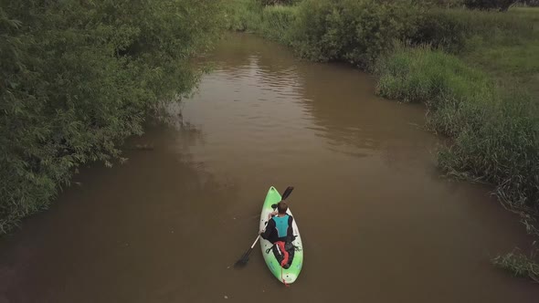 Man kayaking on meandering river with reeds bush’s and trees on either bank. Drone follows behind.