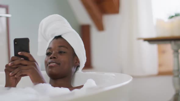 Smiling african american attractive woman relaxing in bath and using smartphone in bathroom