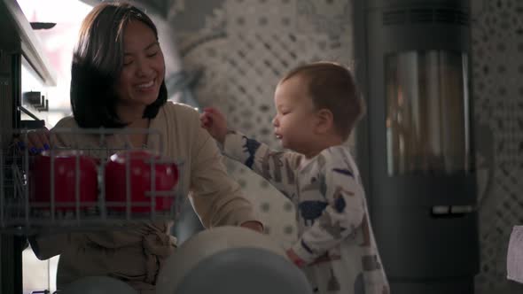 Asian positive son closing the dishwasher with dishes next to mom