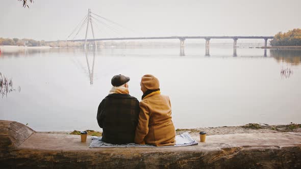 Aged Spouses Talking and Smiling Sitting on Bench at Bank of a River and Looking at Bridge Crossing