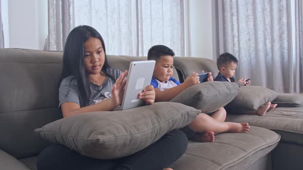 Asian Kids Sitting With Tablet And Mobile Devices On Sofa