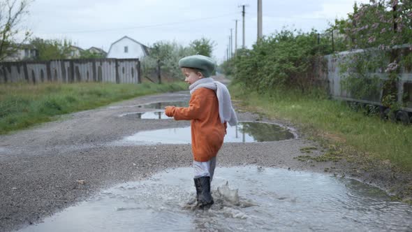 Funny Games, Attractive Cheerful Boy in a Hat and Rubber Boots Plays in Puddles on the Street After