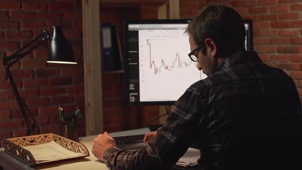 Back View of Caucasian Male Sitting Looking at Monitor with Charts in Light of Table Lamp in