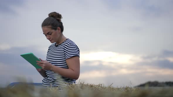 Summer Sky at Sunset with Young Woman Sitting on Haystack on the Left Surfing Social Media on Tablet