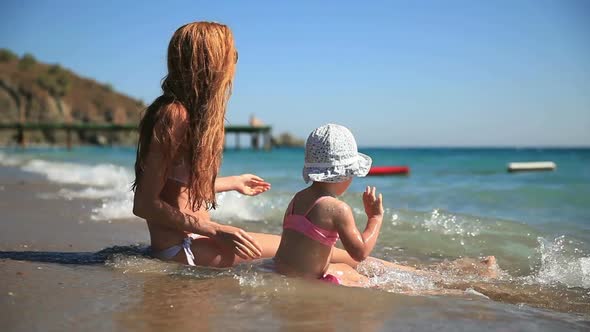 Young Mother with Her Daughter Sitting and Playing in the Sea Waves on the Beach