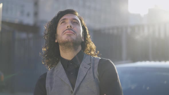 Young Caucasian Man with Green Eyes and Long Hair Looking Up at Sunshine Standing in Urban City