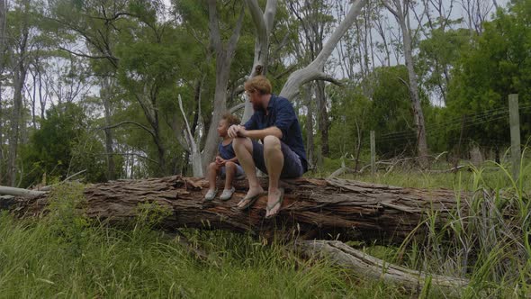 Wide shot of a father and son sitting on a log in the Australian bush looking around.