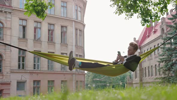 Redhead Man in Suit Relaxing in Hammock and Using Mobile