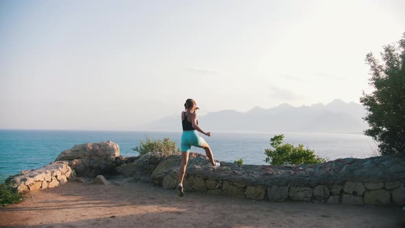 Young Woman in Sports Clothes Running to the Rocky Fence on the Hill By the Sea