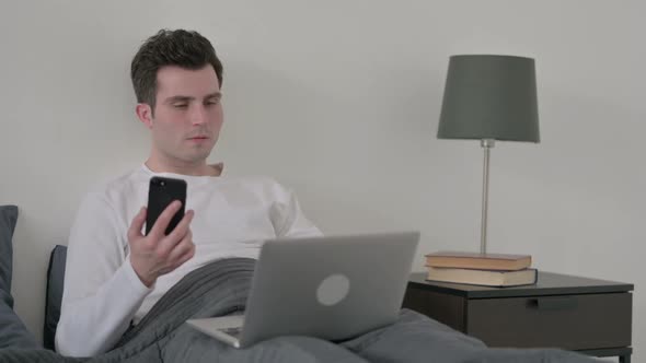 Man with Laptop Using Smartphone in Bed