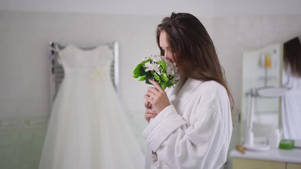 Side View of Young Caucasian Bride Smelling Bridal Bouquet Admiring Wedding Dress Standing in