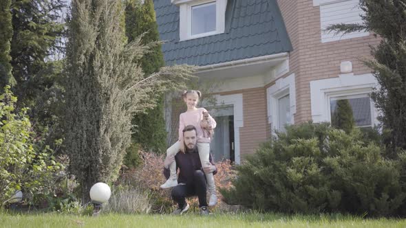 Young Handsome Father Standing in Front of Big House with the Daughter Sitting on His Shoulders. The