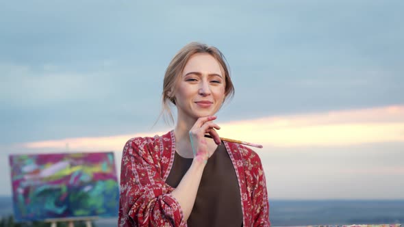 Pretty Young Girl Artist On Sky Background. Behind Her Picture. Girl Touches Her Chin.