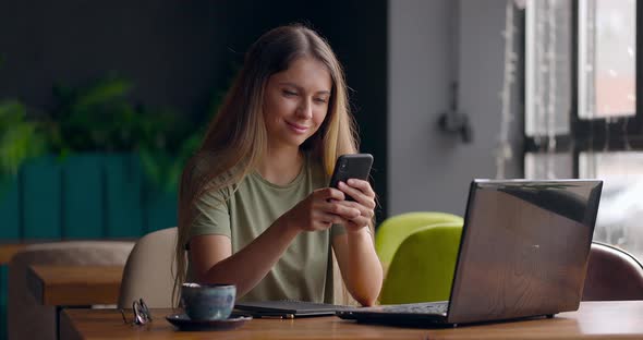 Pretty Woman Sending Message Using Mobile Phone and Smiling While Working with Laptop in Cafe