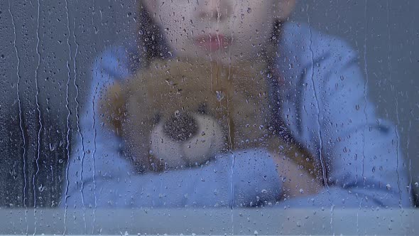 Little Female Orphan Sitting Behind Rainy Window Hugging Teddy Bear and Dreaming