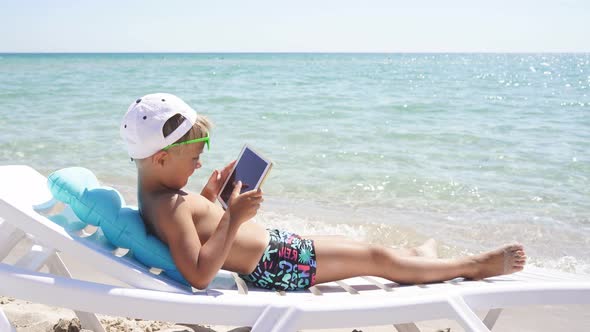 Blonde Child Sitting on the Beach on a Sunny Summer Day Playing on a Tablet