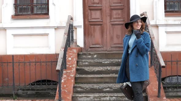 Young Redhaired Woman in a Blue Coat and Black Hat with a Suitcase on the Background of the Old City