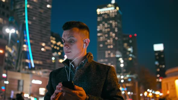 Man with Earphones Is Listening To Music on Phone in City at Night with Lights