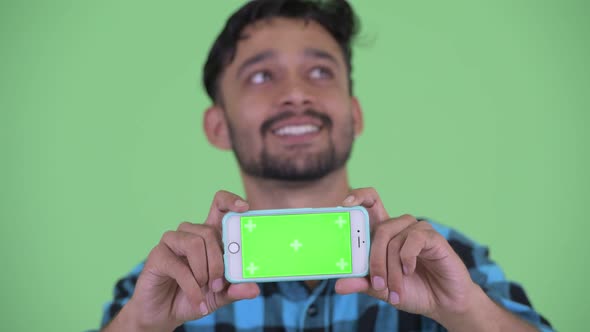 Face of Happy Young Bearded Persian Hipster Man Thinking While Showing Phone