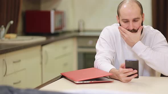 Handsome millennial single guy sit at table in kitchen hold smart phone