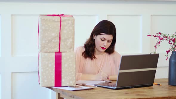 Pretty Young Lady Sitting at Desk and Typing on Laptop and Talking with Present Boxes Icons