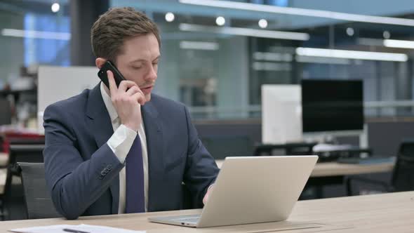 Businessman Talking on Smartphone While Using Laptop in Office